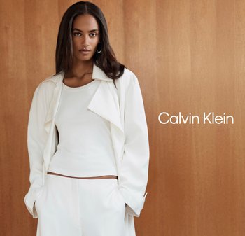 Donna con top bianco senza maniche, giacca bianca ampia e pantaloni coordinati in piedi davanti a uno sfondo in legno con il testo Calvin Klein.