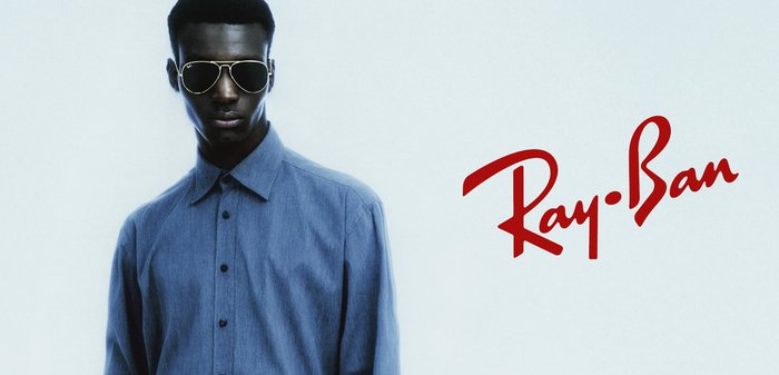 Mężczyzna noszący ciemne okulary przeciwsłoneczne aviator i niebieską koszulę zapinaną na guziki, z czerwonym logo Ray-Ban na dolnej części klatki piersiowej.