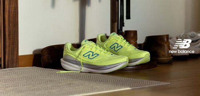 Scarpe da corsa verde lime con dettagli blu, tomaia in mesh, suola ammortizzata e lacci piatti. Posizionate vicino a stivali marroni su pavimento in legno.