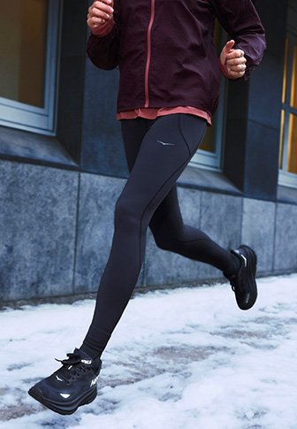 Oseba, ki teče po sneženi pločniku, obuta v črne tekaške čevlje, črne leggins hlače in temno bordo jakno, blizu stavbe.