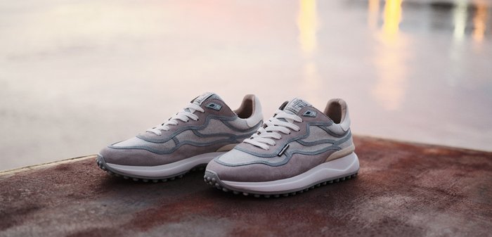 Par grå og hvide afslappede sneakers med teksturerede såler placeret på en stenoverflade ved en rolig vandområde ved solnedgang.