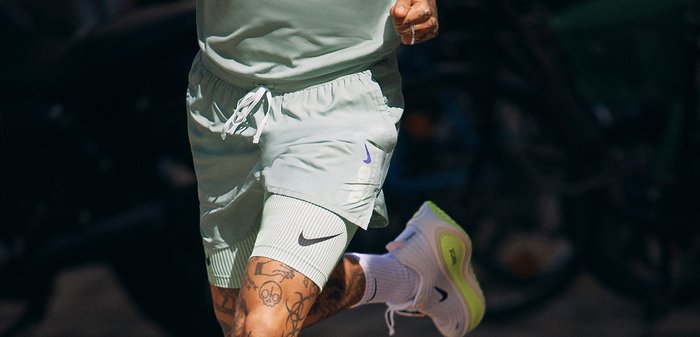 Personne courant à l'extérieur, portant un short vert clair, des chaussures de course blanches avec des semelles vertes et des chaussettes blanches ; tatouages visibles sur les jambes.