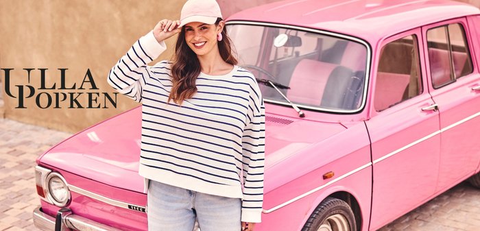 Smiling woman in striped sweater, pink cap, and earrings stands by a vintage pink car on a cobblestone street.