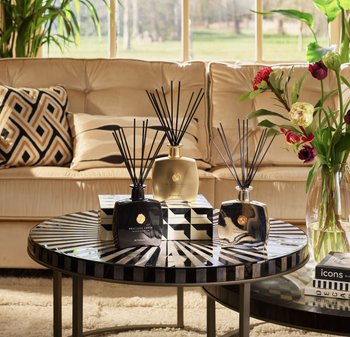 Rund kaffebord med dekorative reed diffusorer, mønstret boks, blomster i vase, bøker og en beige sofa med mønstrede puter i bakgrunnen.