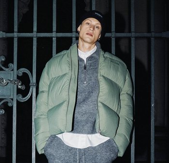 Jasnozielona, puchowa kurtka o pudełkowym kroju, z suwakiem i prążkowanymi detalami, noszona na szarym swetrze i białej koszuli.