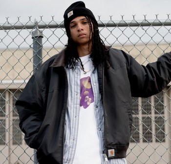 Persona joven con cabello largo trenzado, llevando un gorro negro y una chaqueta bomber, de pie contra una cerca de cadena al aire libre.