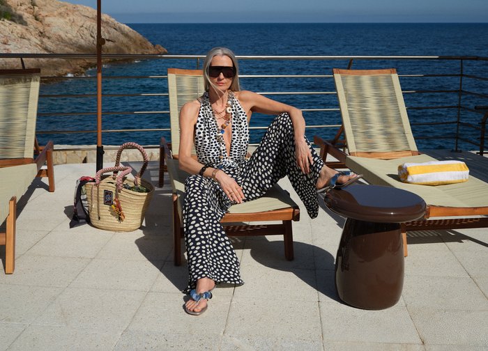 Femme en tenue à pois noirs et blancs assise sur une chaise longue au bord de la mer avec des sacs en osier à proximité et une petite table marron.