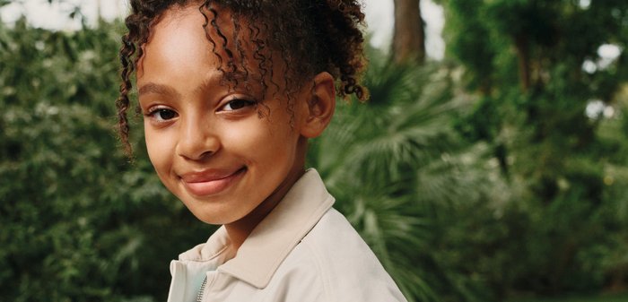 Lächendes Kind mit lockigem Haar, das eine beige Jacke trägt und im Freien vor grünem Baum- und Gras-Hintergrund steht.
