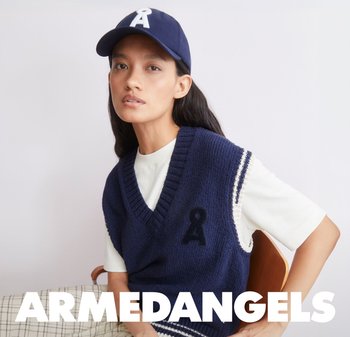 Giovane donna che indossa un gilet lavorato a maglia blu navy e un berretto abbinato con logo bianco, seduta contro uno sfondo chiaro e uniforme.