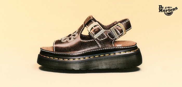 Braune Ledersandale mit blumenförmigem Ausschnitt und gelber Naht, mit Dr. Martens-Logo auf beigem Hintergrund geprägt.