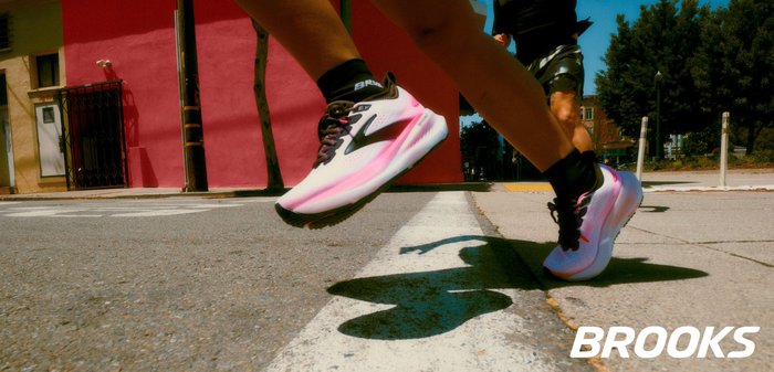 Scarpe da corsa con base bianca, accenti rosa e suola testurizzata, caratterizzate da un design aerodinamico e stringhe nere. Marchio Brooks visibile.