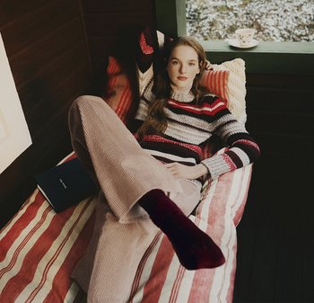 Ung kvinna liggande på randig säng med randig tröja, beige byxor och mörka strumpor, bok i närheten, kopp på fönsterbrädan, lugn inomhusmiljö.