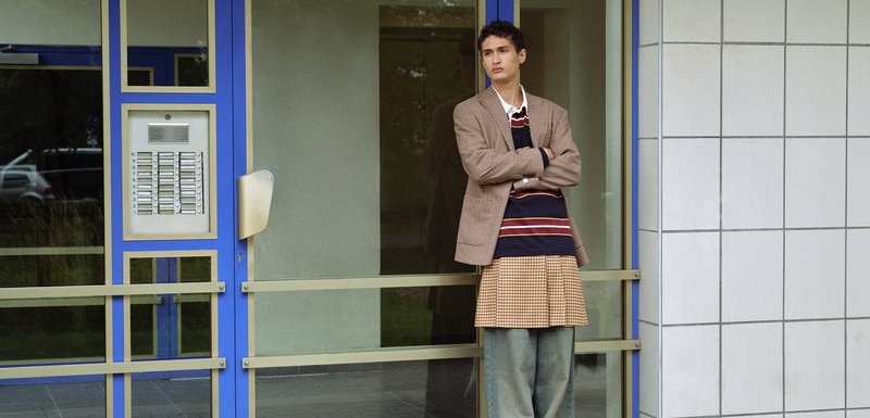 Blazer à carreaux, pull rayé bleu marine, jupe à carreaux et pantalon en denim ample. Se tenant près d'une porte en verre avec un cadre bleu et beige.