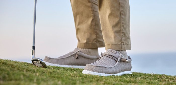 Person iført beige bukser og grå afslappede sko står på græs og holder en golfkølle nær en golfbold.