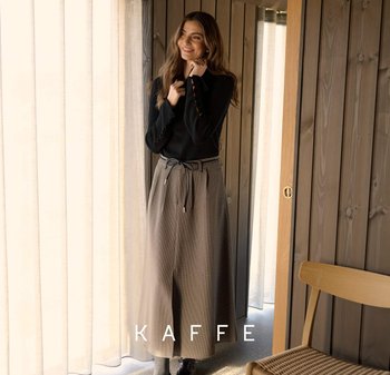 Vrouw die naast een transparante gordijn en een houten muur staat, gekleed in een zwart vest en een beige gemusterde midi-rok met een koordjesriem.