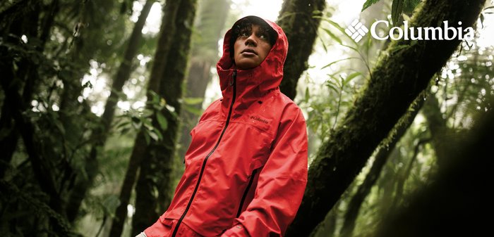 Person wearing a red Columbia rain jacket with hood up, standing in a misty forest surrounded by moss-covered trees and green foliage.