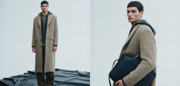 Beiger Wollmantel mit großen Revers, darüber ein dunkler grüner Kapuzenpullover mit Reißverschluss und ein gestreiftes Hemd, kombiniert mit braunen Hosen und einer schwarzen Tasche.