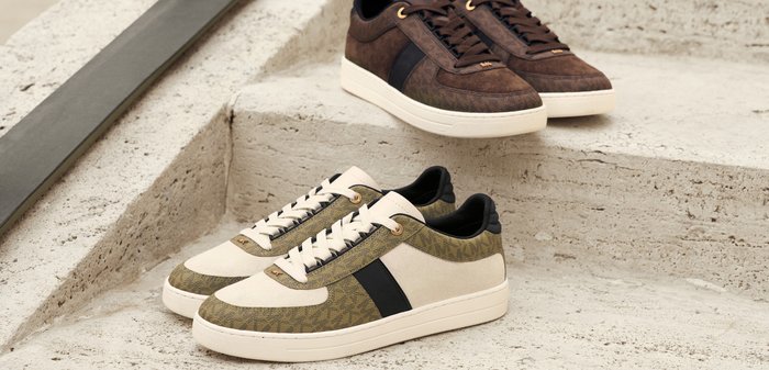 Due paia di sneakers su una superficie strutturata. Un paio ha un design in suede marrone con accenti neri; l'altro è color crema con motivi verde oliva.