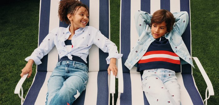 Zwei Kinder entspannen auf blau-weiß gestreiften Stühlen im Freien, lächeln und relaxen, bekleidet mit lässigen Jeans und leichten Jacken.