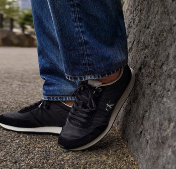 Zwarte sportieve schoenen van suede en mesh, met veters, een witte zool en een klein logo. Gepaarde met blauwe denim jeans.