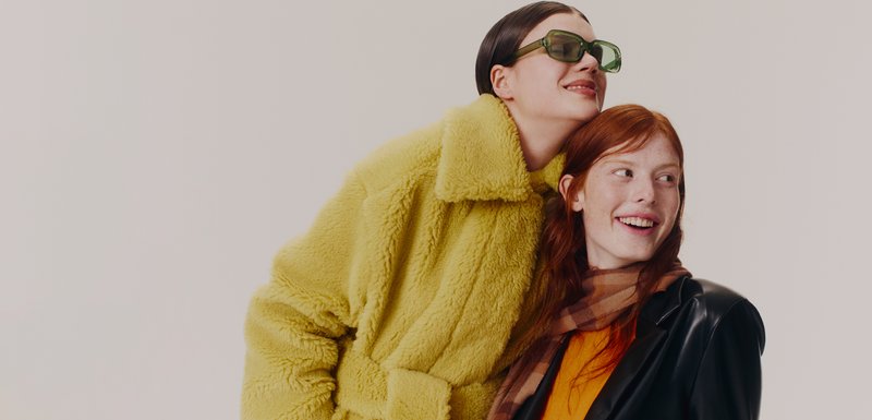 Deux mannequins posent côte à côte. L'un porte un manteau jaune duveteux et des lunettes vertes ; l'autre a une veste en cuir noire et une écharpe à carreaux.