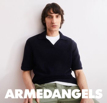 Giovane uomo con capelli scuri di media lunghezza, che indossa un maglione nero a maniche corte e pantaloni color oliva, seduto contro un muro bianco, con la scritta "ARMEDANGELS" sotto.