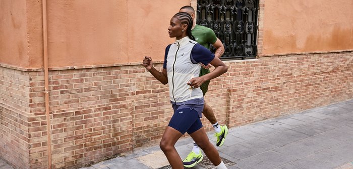 Δύο δρομείς, μια γυναίκα με σορτς και αμάνικο τοπ και ένας άνδρας με πράσινο μπλουζάκι και σορτς, τρέχουν σε πεζοδρόμιο της πόλης δίπλα σε τοίχο από τούβλα και σοβά.