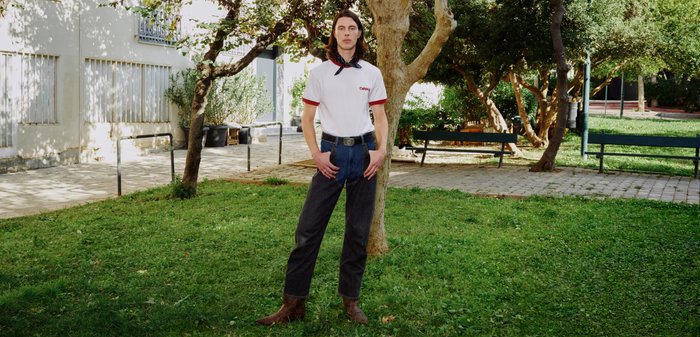 Νεαρό άτομο με μακριά μαλλιά στέκεται στο γρασίδι κοντά σε δέντρα, φορώντας λευκό μπλουζάκι, σκούρο παντελόνι, καφέ μπότες και κασκόλ στο λαιμό σε ένα ηλιόλουστο πάρκο.