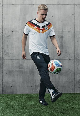 Biała koszulka piłkarska z czerwoną i pomarańczową wzór w romby przy kołnierzu, połączona z czarnymi spodniami i czarnym obuwiem, stojąca na trawie.