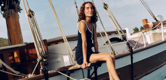 Young woman in black dress sits barefoot on a wooden boat ladder above calm water on a sunny day with ropes and sails visible.