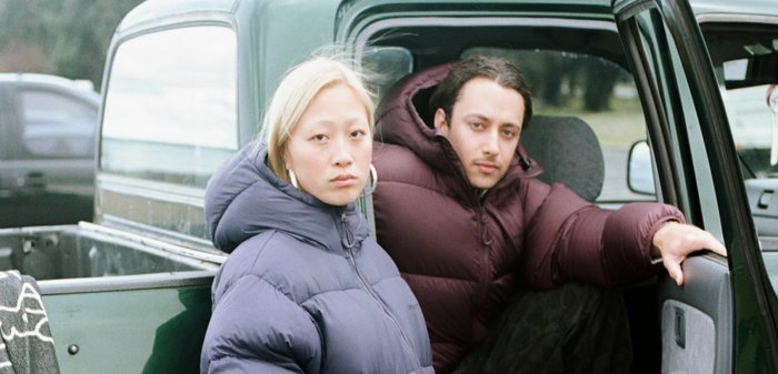 Dos personas están sentadas en la caja de una camioneta. Una lleva una chaqueta acolchada azul y la otra una chaqueta acolchada burdeos, ambas con capucha.