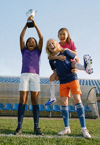Drie meisjes vieren het op een voetbalveld; één houdt een trofee vast, een ander draagt een teamgenoot op haar rug, allemaal lachen ze vrolijk.