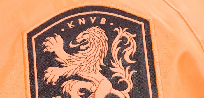 Μαύρο και πορτοκαλί σήμα KNVB με λεπτομερή σχεδιασμό λιονταριού σε πορτοκαλί ύφασμα φόντου.