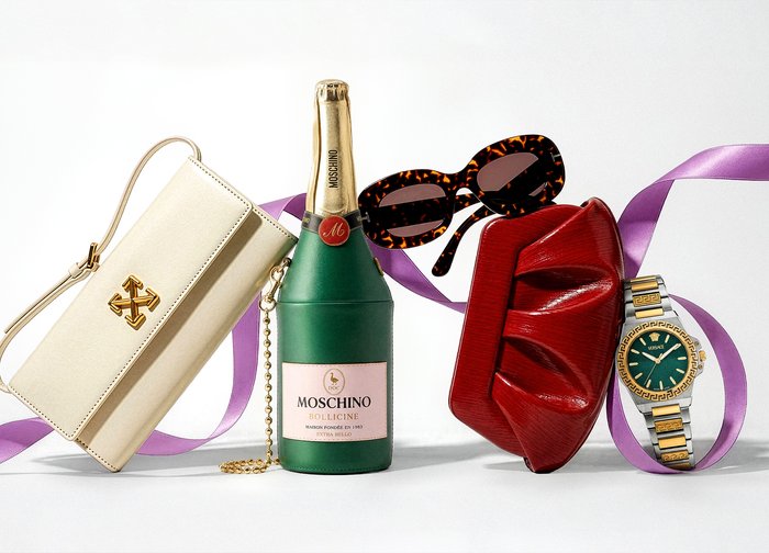 Λευκή τσάντα σχεδιαστή, πράσινο μπουκάλι σαμπάνιας Moschino, γυαλιά ηλίου με καφέ σχέδιο χελώνας, κόκκινο φάκελος και χρυσό ρολόι Versace με μοβ κορδέλα.