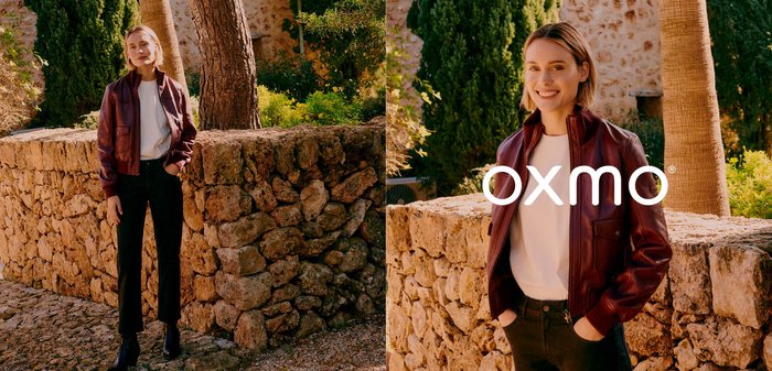 Junge Frau lächelt mit den Händen in den Taschen, trägt eine braune Lederjacke und ein weißes Hemd, steht draußen an einer Steinmauer.