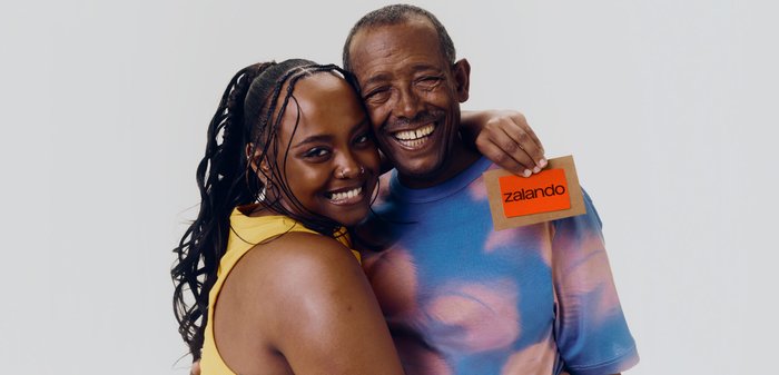 Femme souriante en jaune et homme en chemise tie-dye bleu-rose s’enlacent, l’homme tient une carte orange avec le texte « zalando » sur un fond uni.