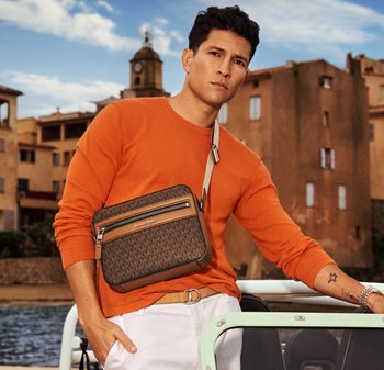 Hombre con suéter naranja y pantalones blancos, llevando un bolso cruzado marrón con patrón, apoyado en la barandilla de un barco con edificios y agua de fondo.