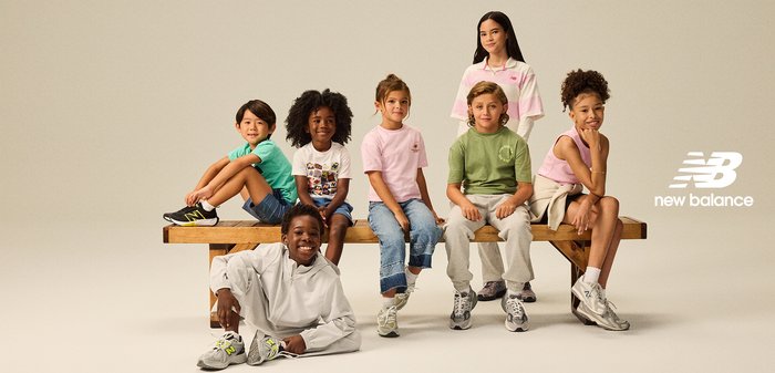 Sete crianças sentam-se num banco de madeira, vestindo roupas casuais de várias cores. Os sapatos incluem ténis pretos, cinzentos e brancos com detalhes.