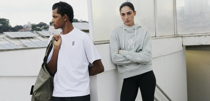 Zwei Personen stehen an einer weißen Dachmauer; eine trägt ein weißes T-Shirt und hält eine grüne Tasche, die andere einen grauen Kapuzenpullover mit verschränkten Armen.