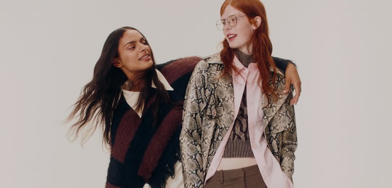 Deux femmes posent ensemble. L'une porte un pull en peluche rayé bordeaux et noir ; l'autre porte une veste imprimée serpent sur une chemise rose et un pantalon marron.