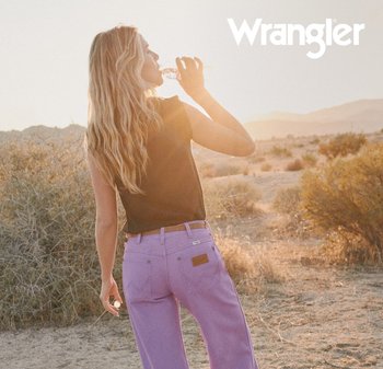 Vrouw met lang blond haar, gekleed in paarse spijkerbroek en een zwart mouwloos topje, drinkt water buiten bij zonsondergang in een droog landschap met struikjes.