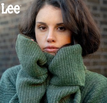 Grön stickad tröja med oversized, vikt krage. Mjuk textur och ribbad mönster. Modellen håller kragen nära ansiktet.