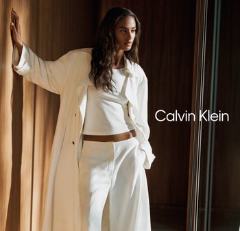 Mujer con camisa blanca holgada, top corto y pantalones, recostada contra una pared de madera en una habitación iluminada suavemente, con el texto "Calvin Klein" a su lado.