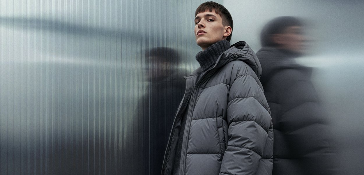 Doudoune grise avec capuche, présentant un design matelassé et une texture lisse. Portée sur un col roulé en tricot foncé, devant un arrière-plan métallique flou.