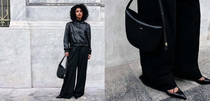 Woman with curly hair wearing a black leather jacket, wide black pants, pointed shoes, holding a black handbag against a stone wall.