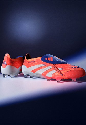 Φωτεινά πορτοκαλί παπούτσια ποδοσφαίρου Adidas Predator με μπλε λεπτομέρειες και λευκές σόλες, τοποθετημένα σε ένα σκούρο γκράντ γκράντ φόντο.