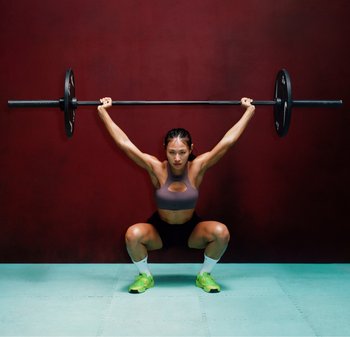 Haltère avec poids soulevé au-dessus de la tête par une personne portant un crop top athlétique gris et un short noir, debout sur un tapis bleu sarcelle contre un mur rouge.