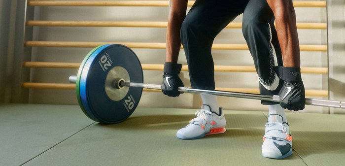 Kesztyűt és sportcipőt viselő személy egy 20 kg-os súlytárcsákkal ellátott rúd súlyt emel egy edzőtermi környezetben.