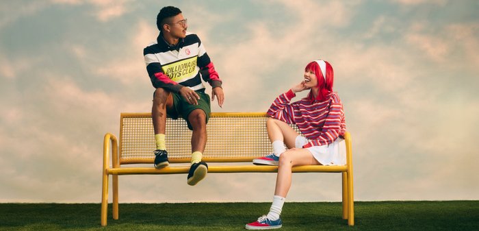Deux jeunes adultes, l'un aux cheveux roux et pull rayé assis sur un banc jaune, l'autre portant des lunettes et un short assis sur le dossier.