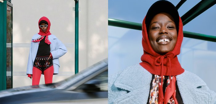 Manteau bleu clair texturé, haut noir, écharpe rouge et short à motifs avec des leggings rouges ; lunettes de soleil surdimensionnées ; souriant devant un fond uni.